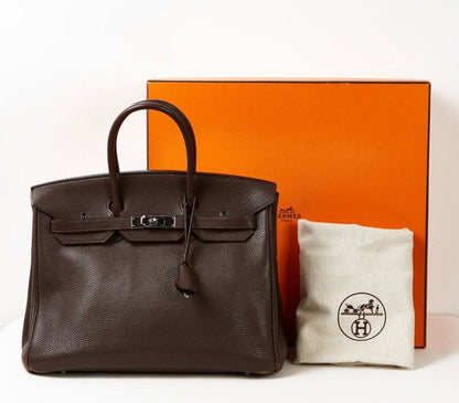 Brown leather handbag with orange box and white pouch on a light gray background